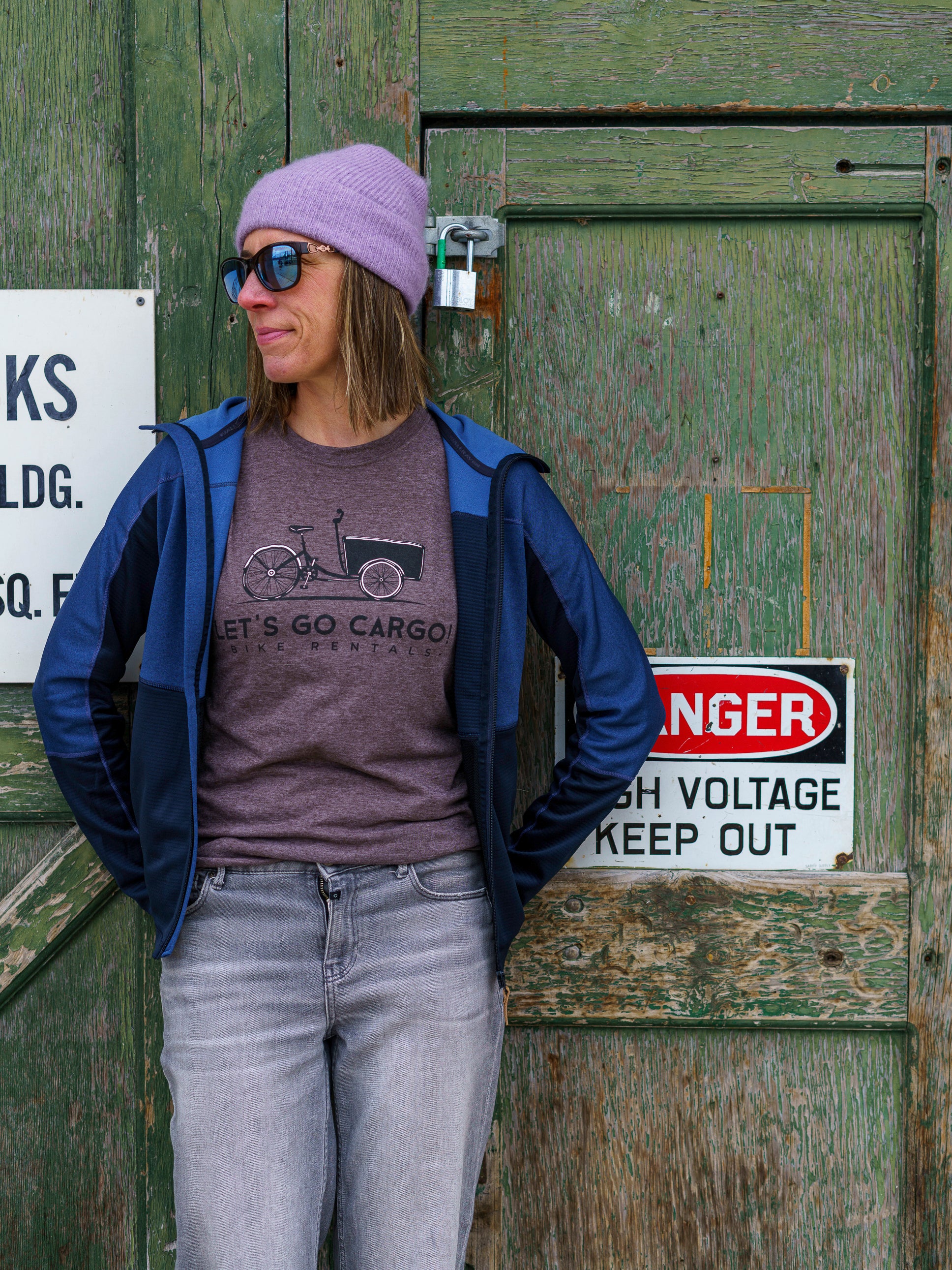 Person wearing a purple touque and sunglasses, standing in front of a wooden door with signs.