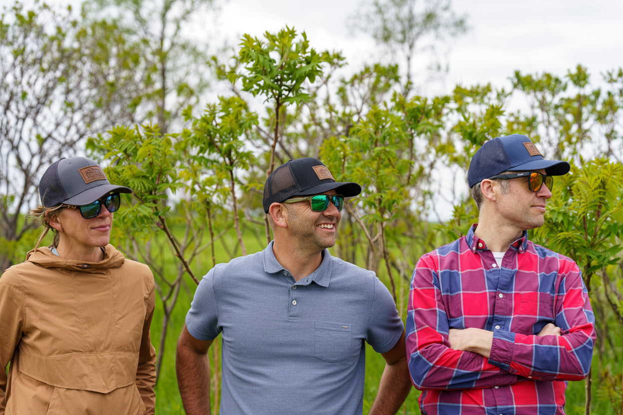 Three people wearing sunglasses and caps outdoors with greenery in the background
