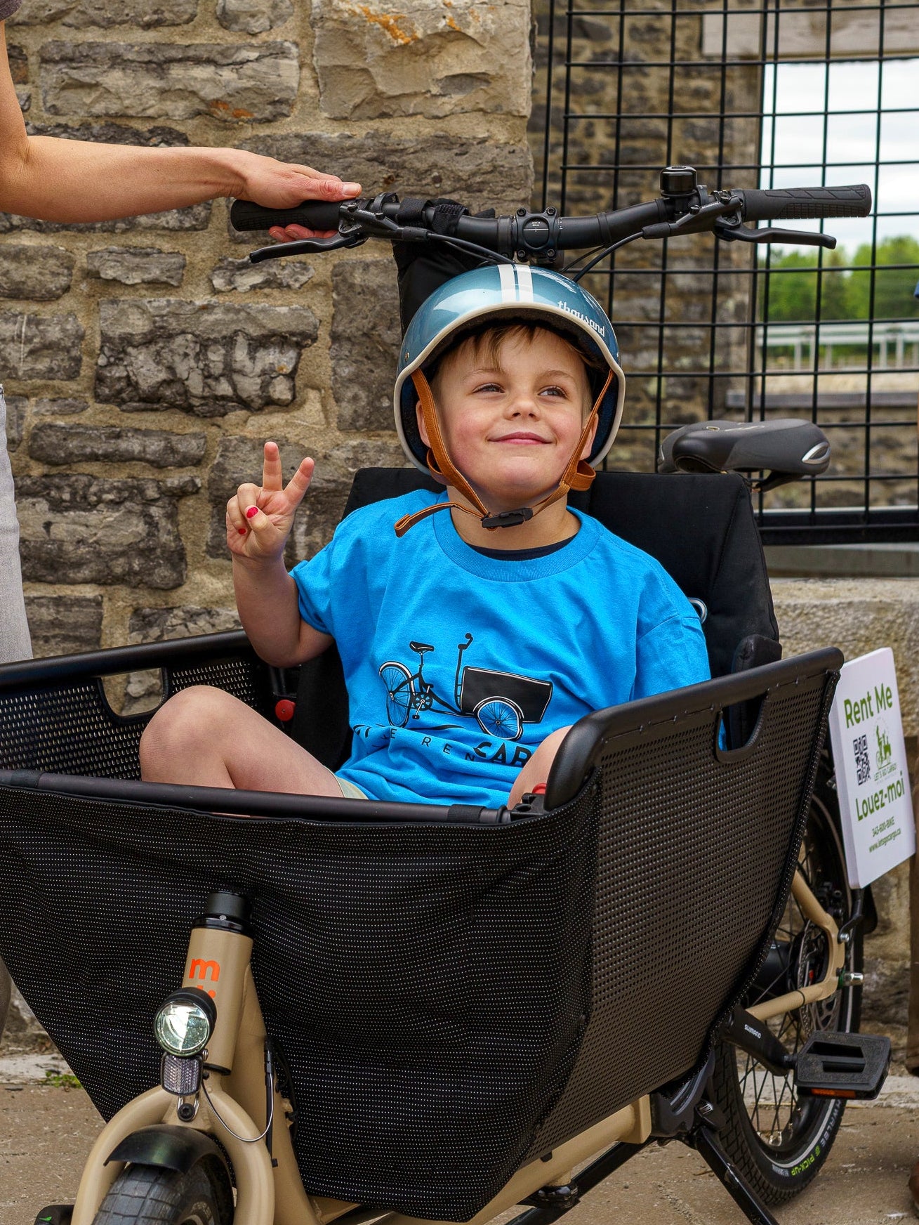 Child in a cargo bike with a helmet, sitting next to a stone wall.